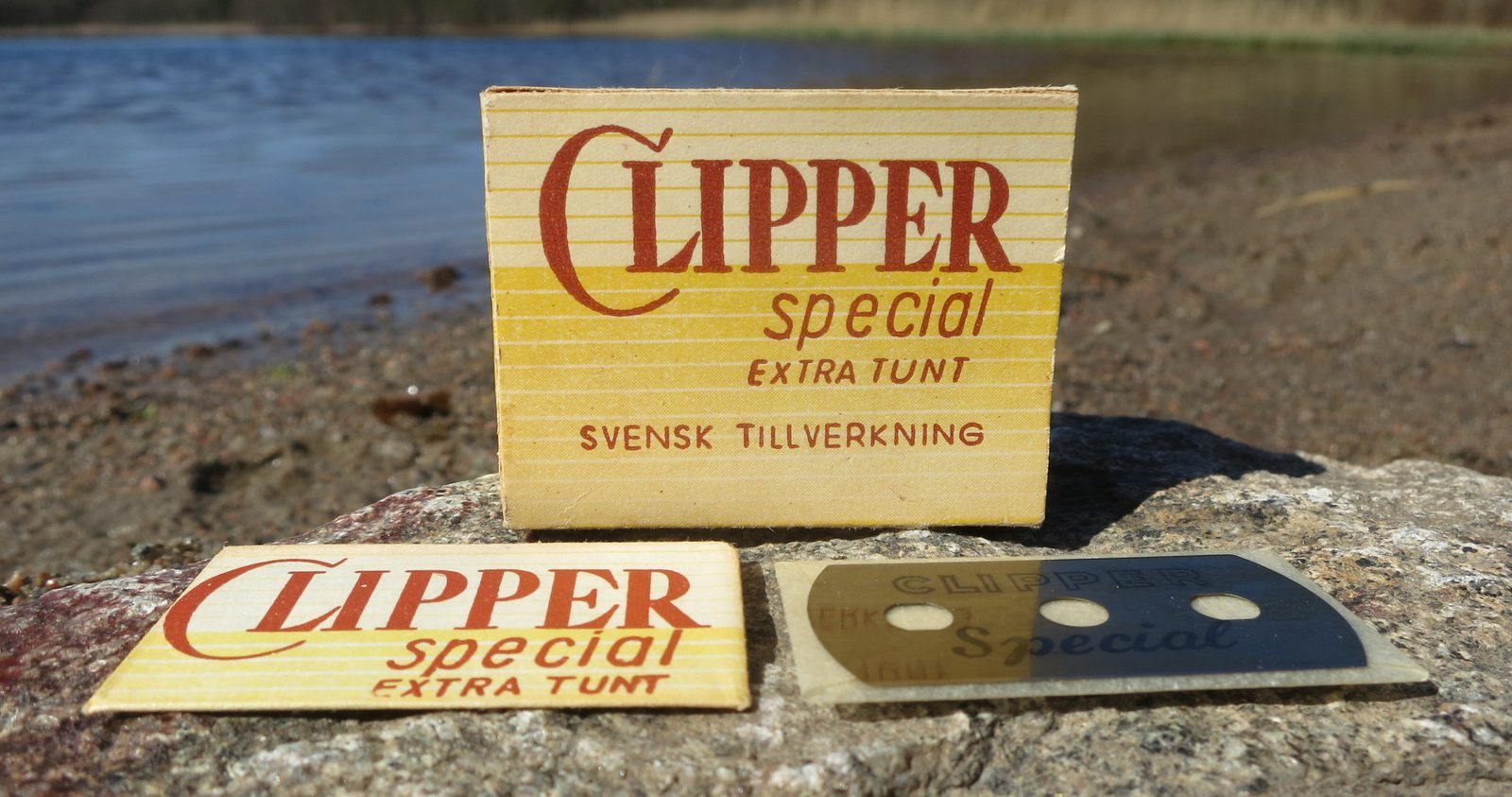 A box of Clipper Special razor blades sits on a rock by water, with a blade and wrapper in front