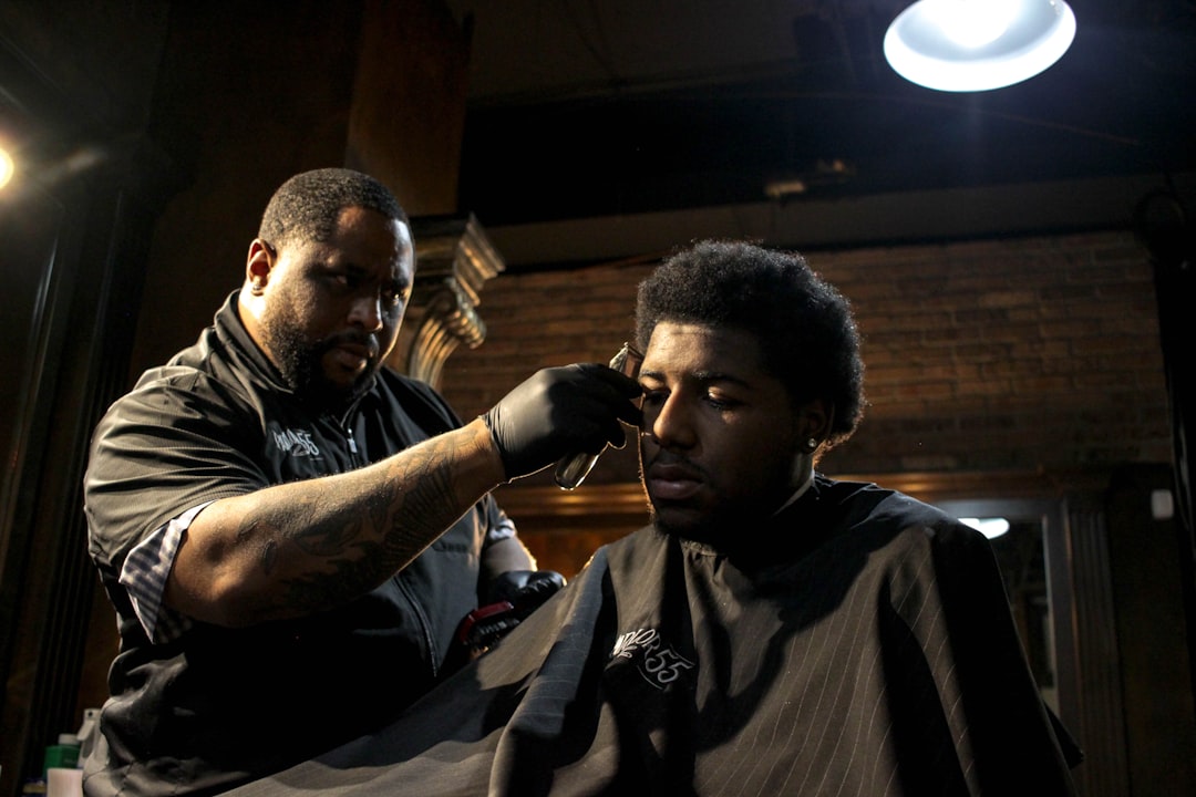 A barber with black gloves uses clippers on a seated man's hair in a dimly lit shop