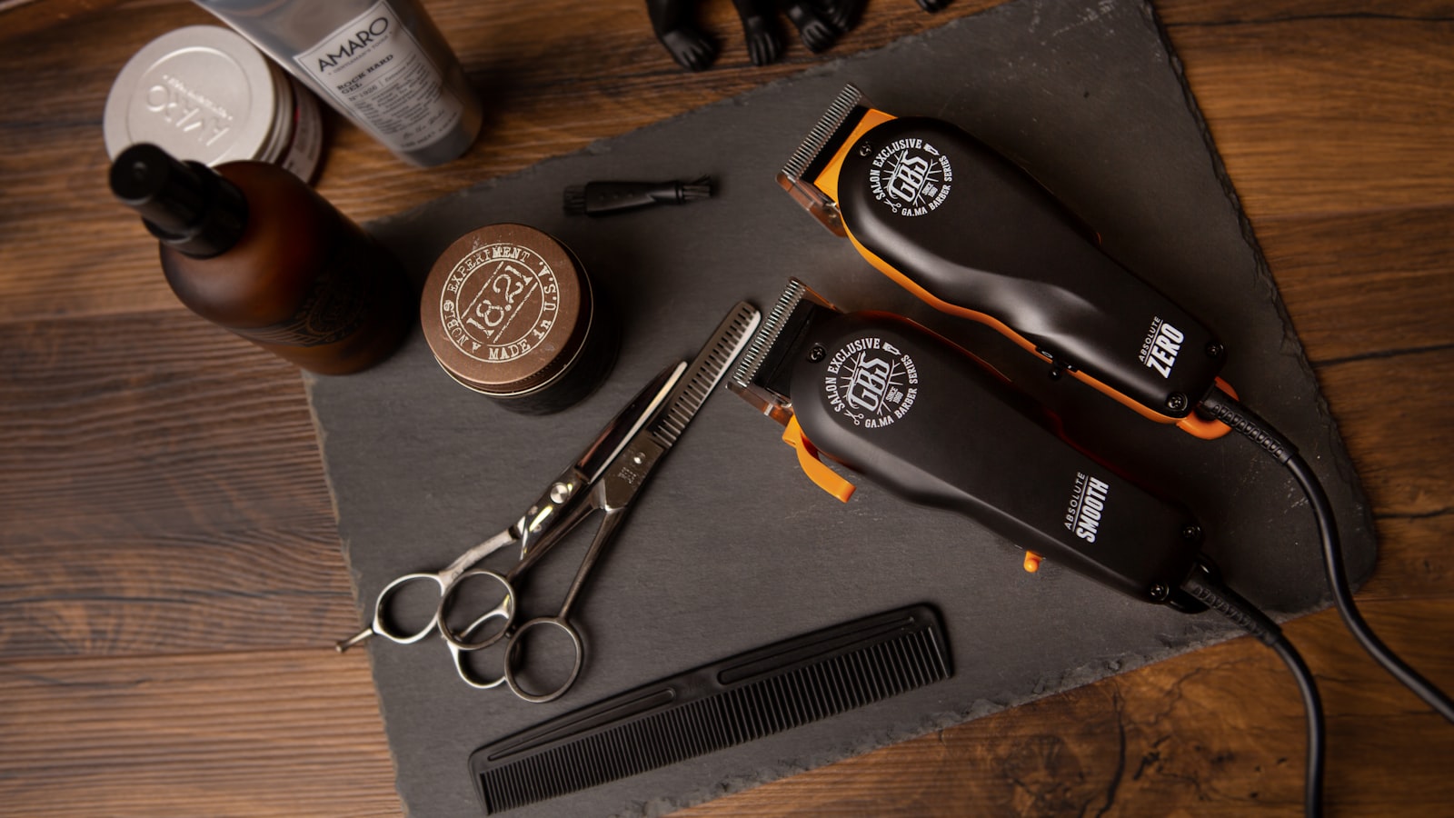 Hair clippers, scissors, comb, jars, and bottles are arranged on a dark slate board