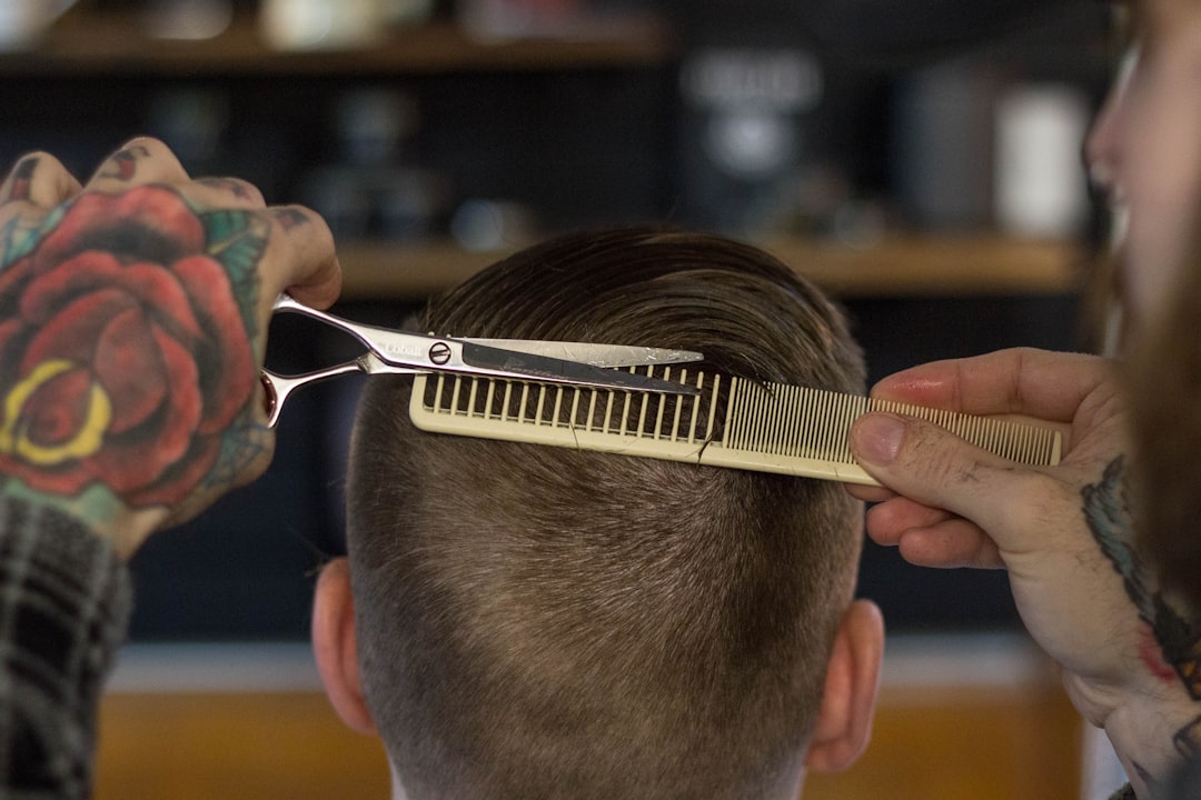A barber with tattooed hands cuts a man's hair using scissors and a comb
