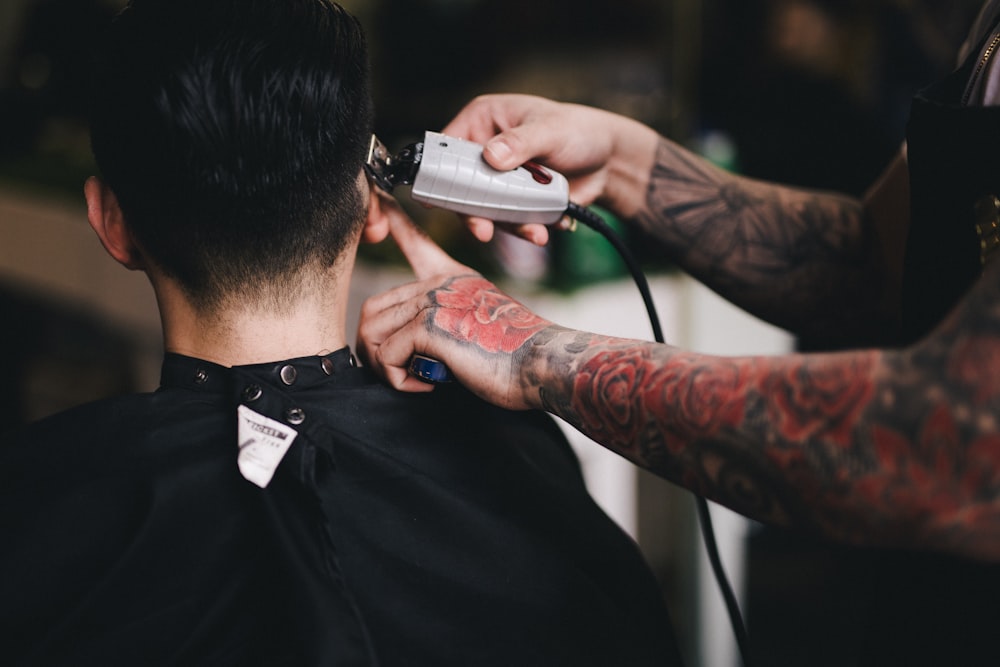 A tattooed barber uses clippers to trim the hair of a client wearing a black cape