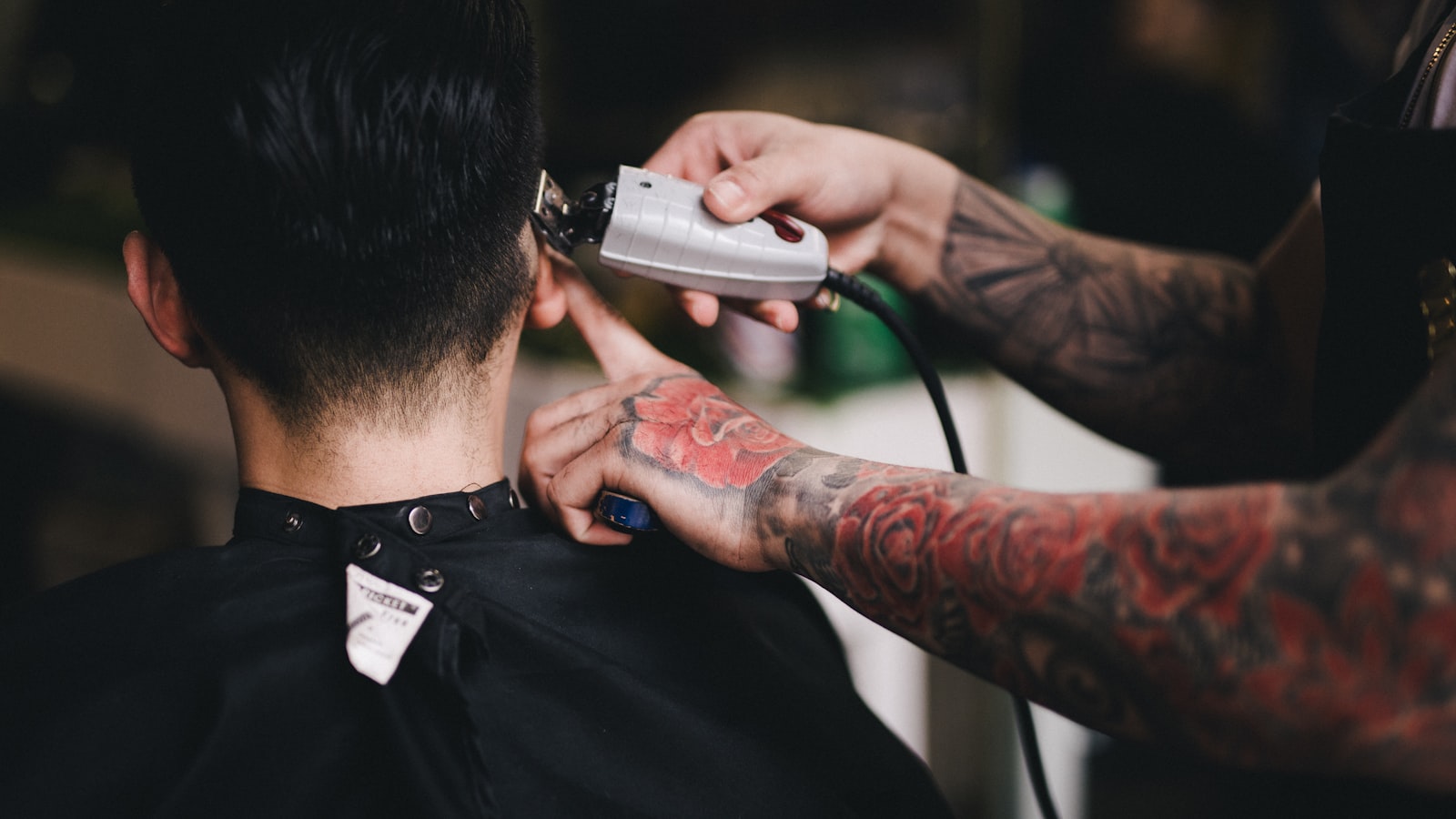 A tattooed barber uses clippers to cut a man's hair in a salon