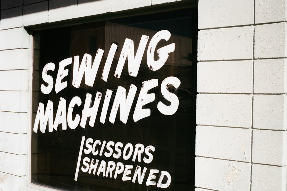 White painted lettering on a dark window reads "SEWING MACHINES SCISSORS SHARPENED" on a white brick wall