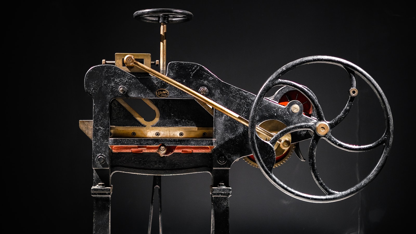 An antique cast iron machine with brass accents, a large side wheel, and top handwheel on a black background